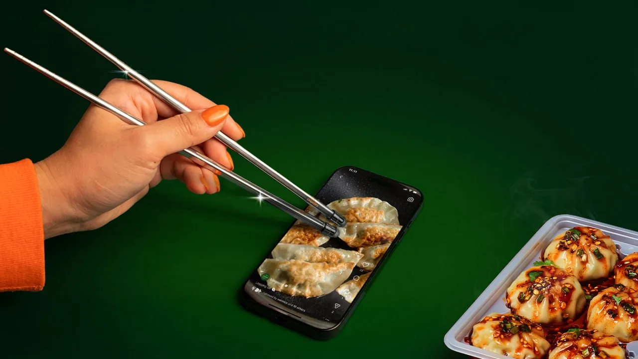 Hand using chopsticks to tap dumplings on a smartphone screen next to a tray of dumplings, suggesting playful digital engagement