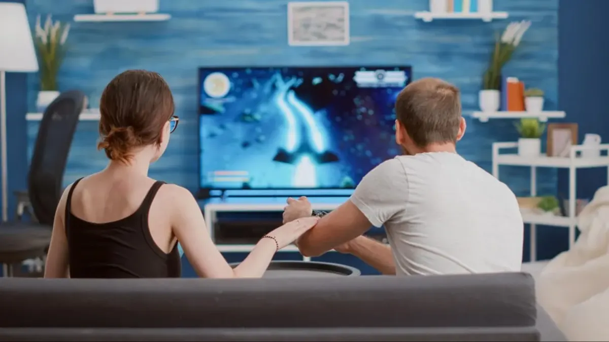 Two people sit on a couch watching a video game on TV, suggesting players experiencing gameplay together at home
