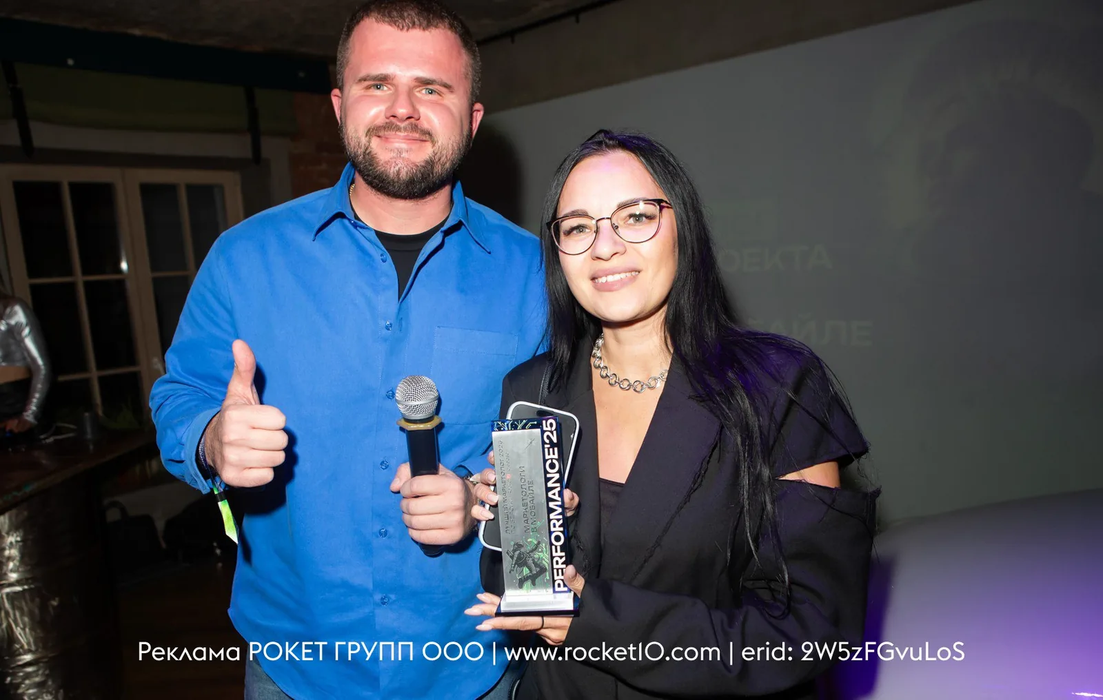 Two people at an event, posing with a microphone and an award in an indoor setting