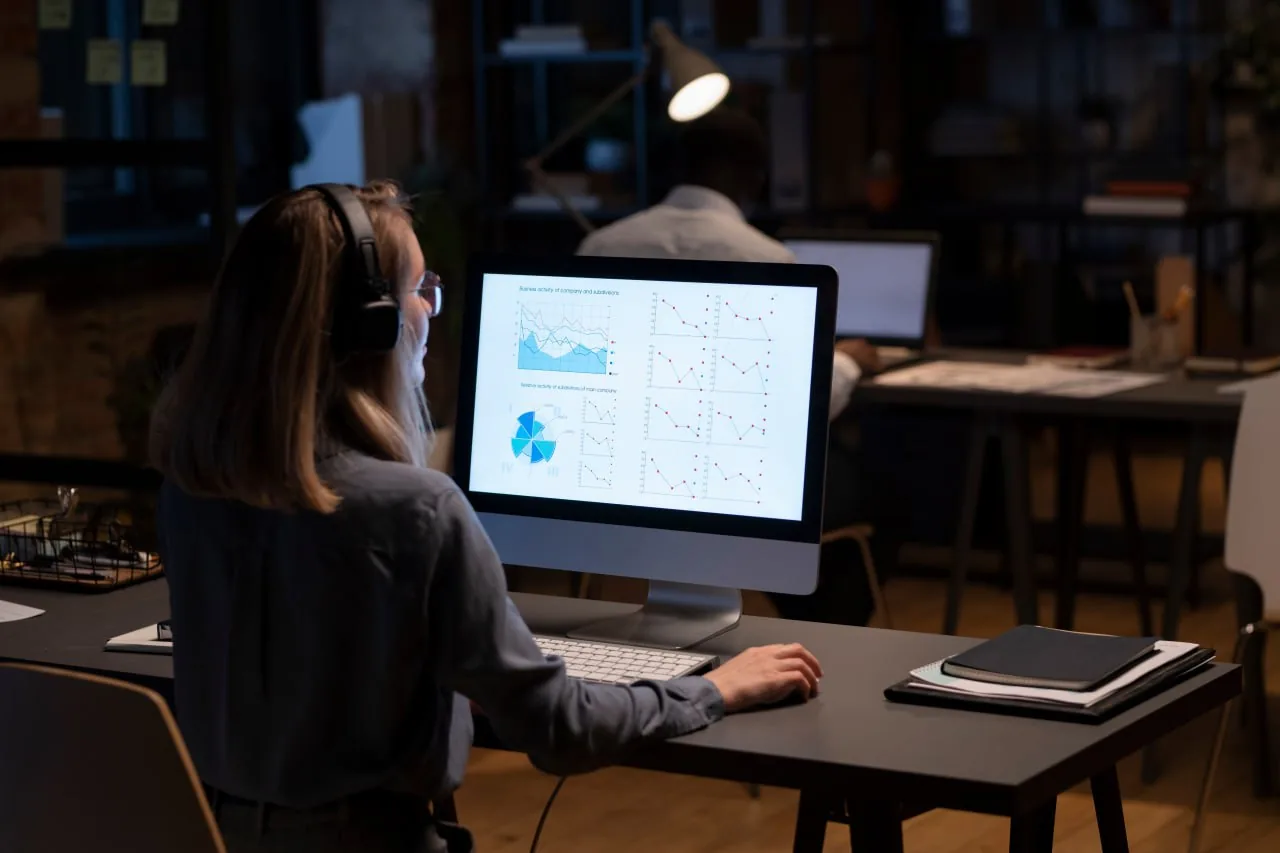 Woman wearing headphones reviewing charts on a desktop monitor in an office