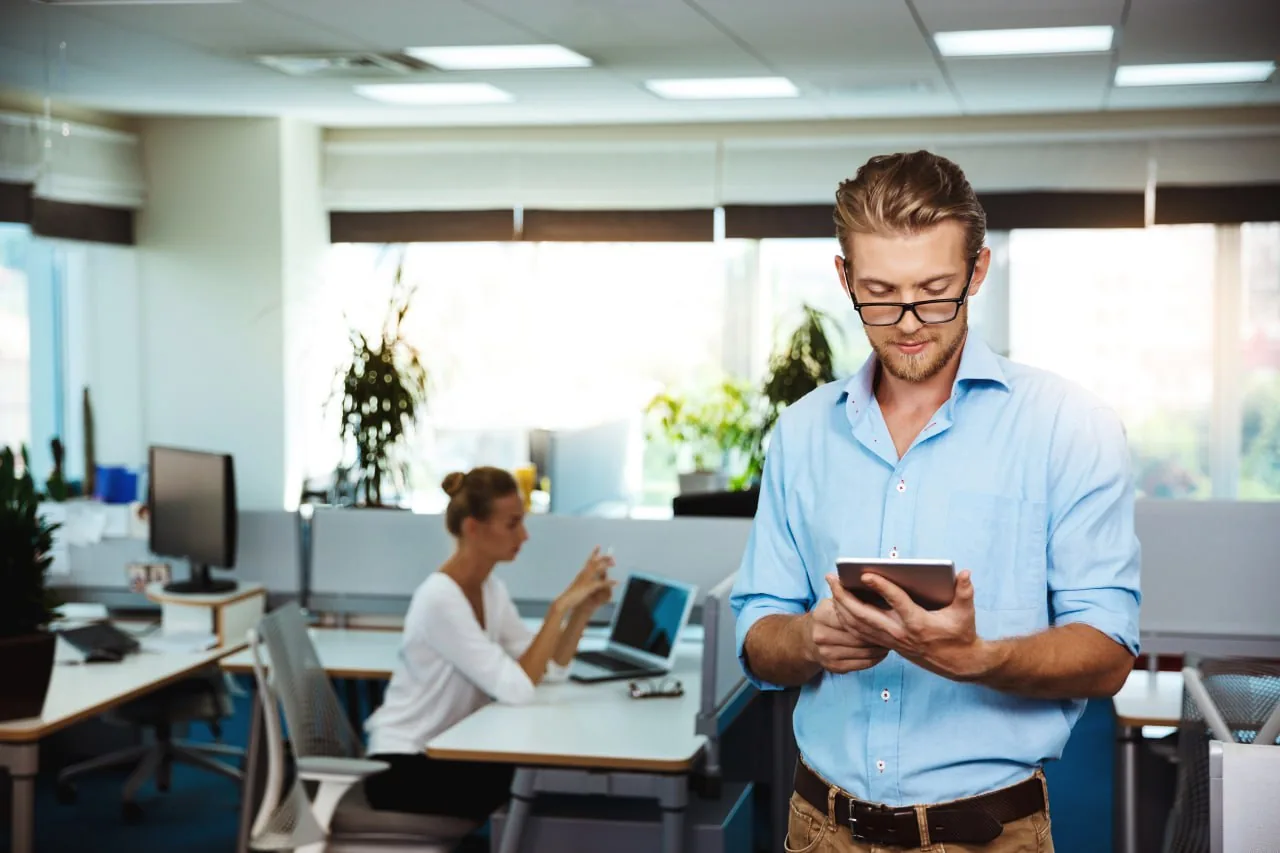 Man in office using smartphone with colleague working on laptop in background