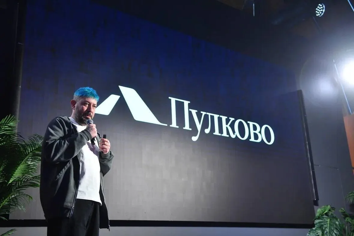 Bearded man with blue hair speaking into a microphone on stage in front of a backdrop reading “Пулково